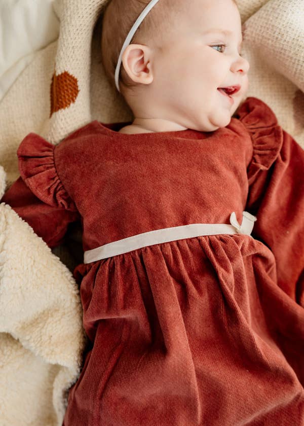 Baby in a rust-colored dress with a white headband, surrounded by soft textures.