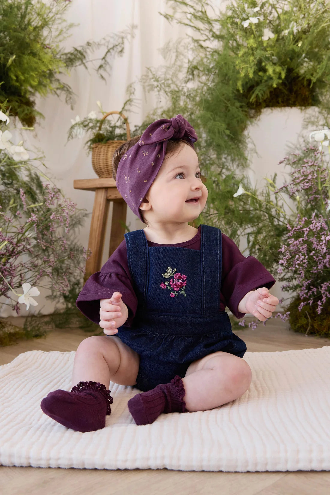 A baby girl sitting on a white textured blanket in a decorated nursery setting, wearing a dark denim overall romper over a deep purple long-sleeve shirt, a matching purple bow headband, and purple booties.