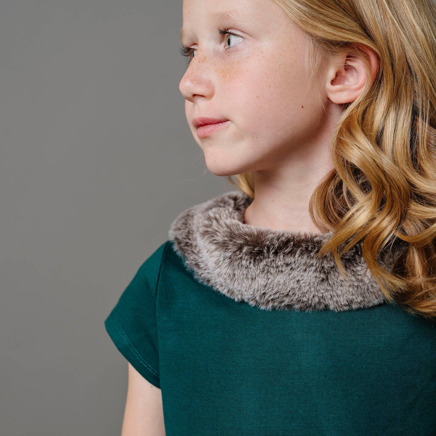 Young girl wearing a teal shirt with a fur collar against a gray background