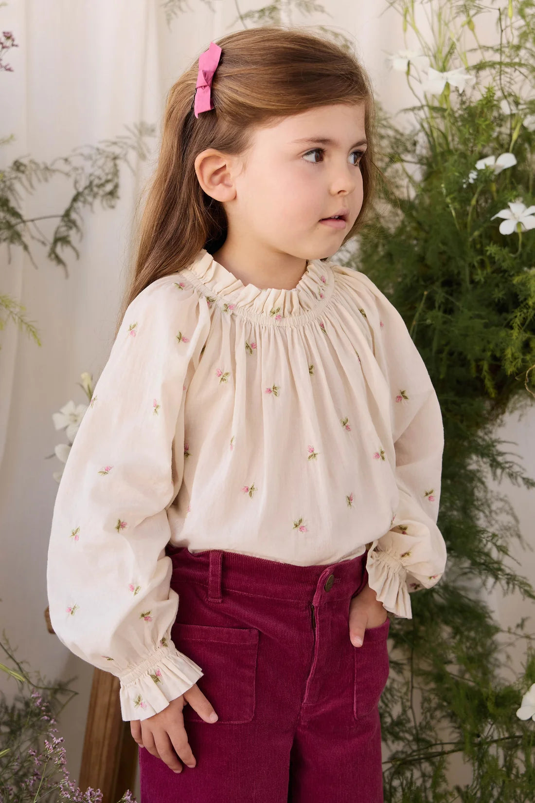 A young girl with light brown hair tied back with a pink bow is wearing a cream-colored long-sleeve blouse with delicate floral embroidery and ruffled details, paired with magenta corduroy pants. She is posed in front of a soft-focus background of green foliage and white flowers.