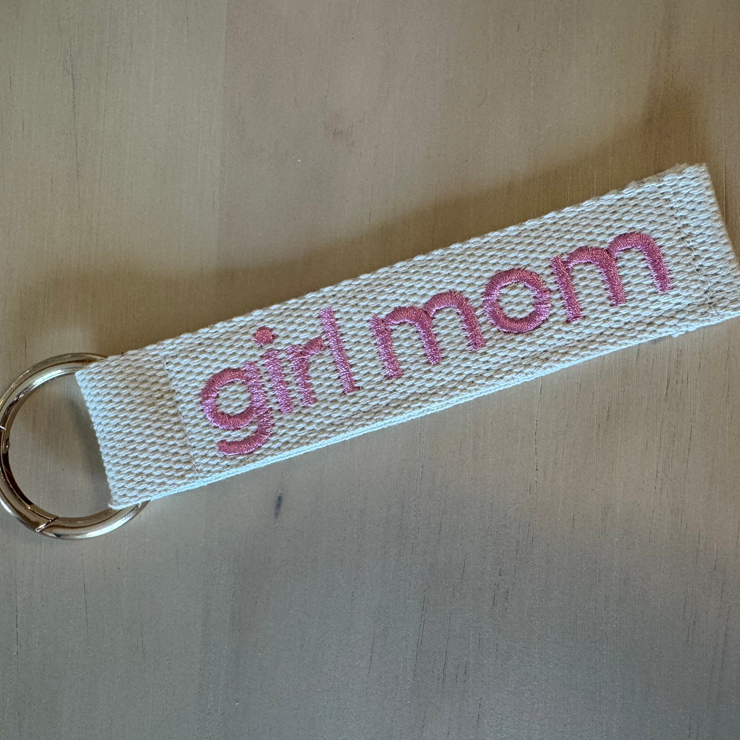 A fabric keychain with a white woven strap featuring the words 'girl mom' in pink embroidered text, attached to a gold metal key ring, shown on a light wood surface.