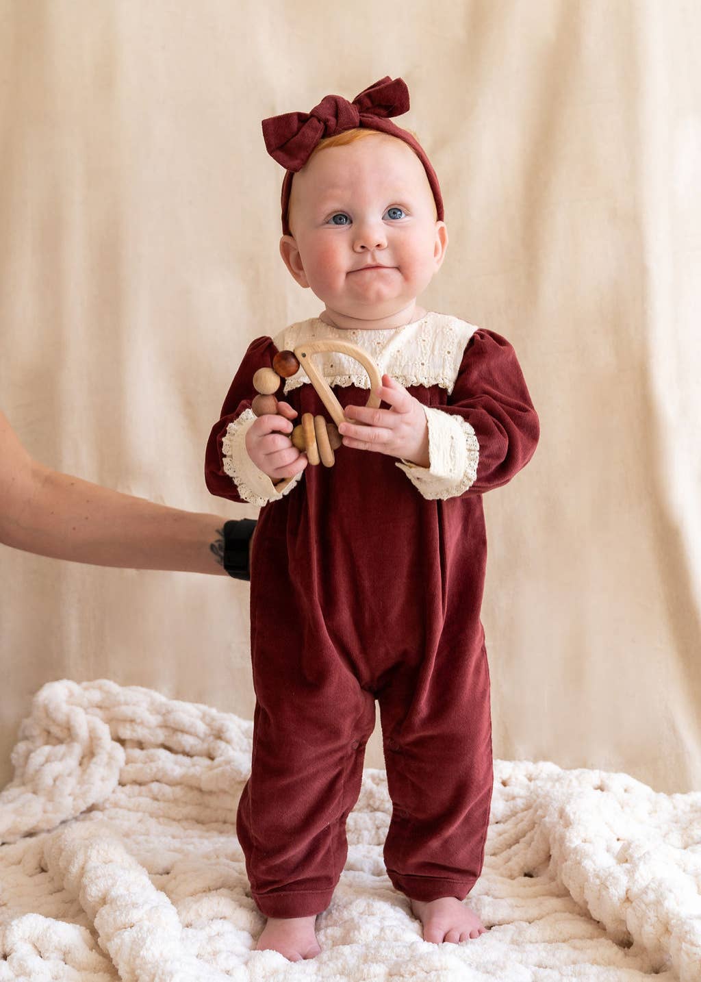 Baby in a burgundy outfit with a bow holding a wooden toy on a beige blanket.