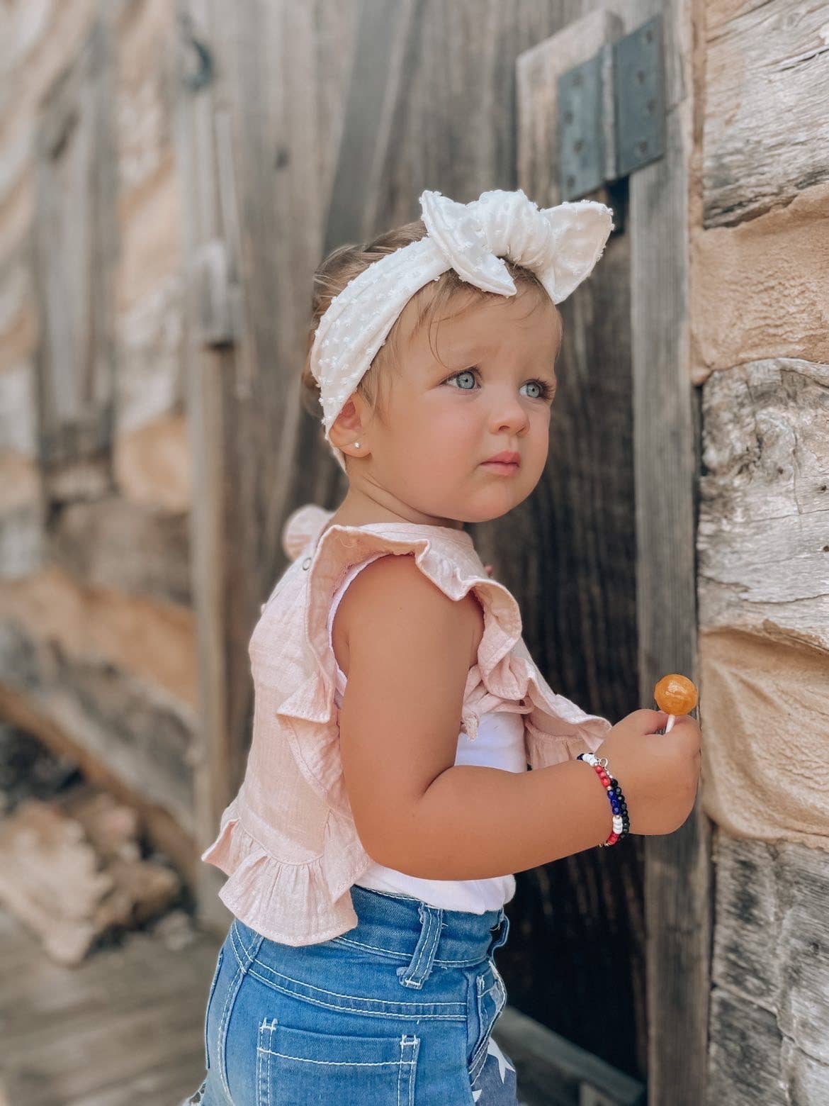 Young girl wearing a pink top and blue jeans with a white headband, standing against a stone wall.
