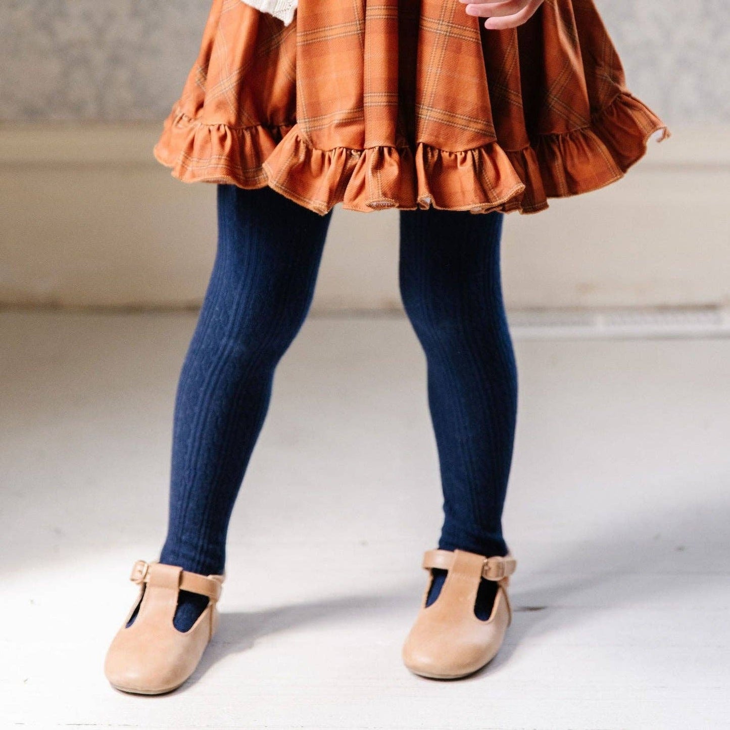 Child wearing a rust-colored skirt, blue leggings, and beige shoes on a neutral background