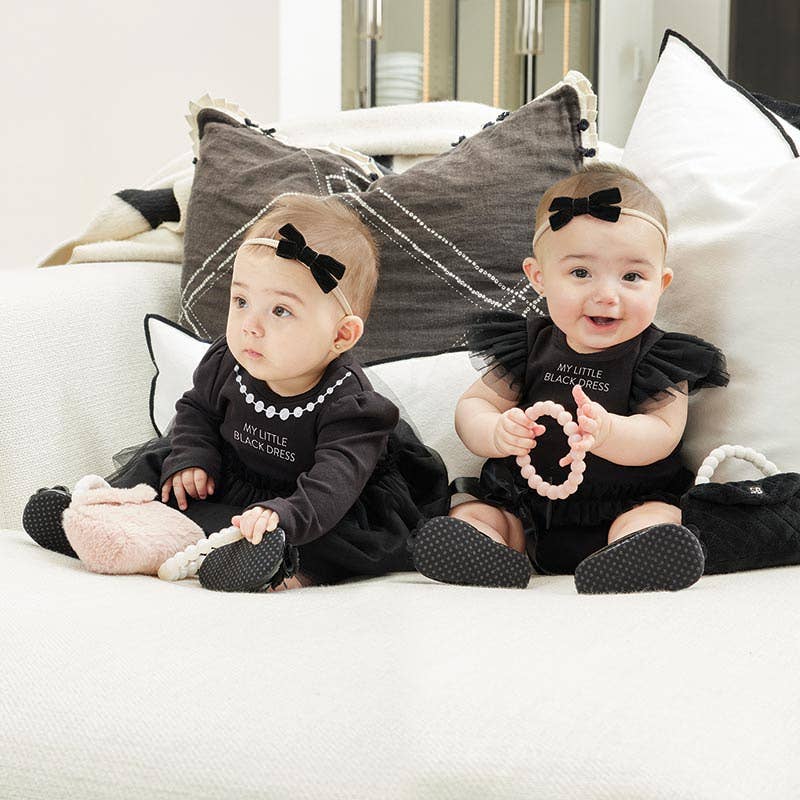 Two babies in matching black outfits with bows sitting on a couch.
