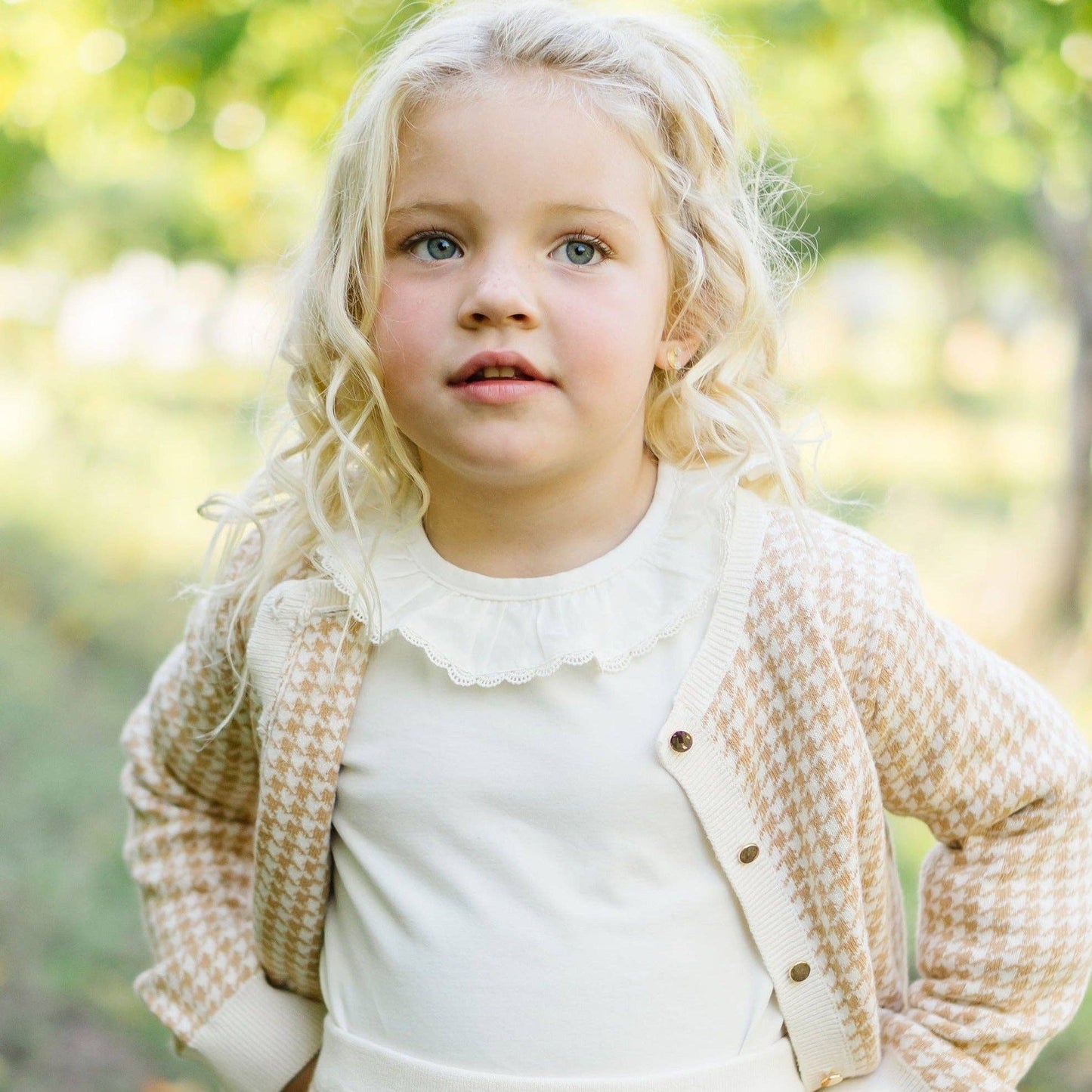 Young girl with blonde hair wearing a beige cardigan over a white shirt in a natural setting.