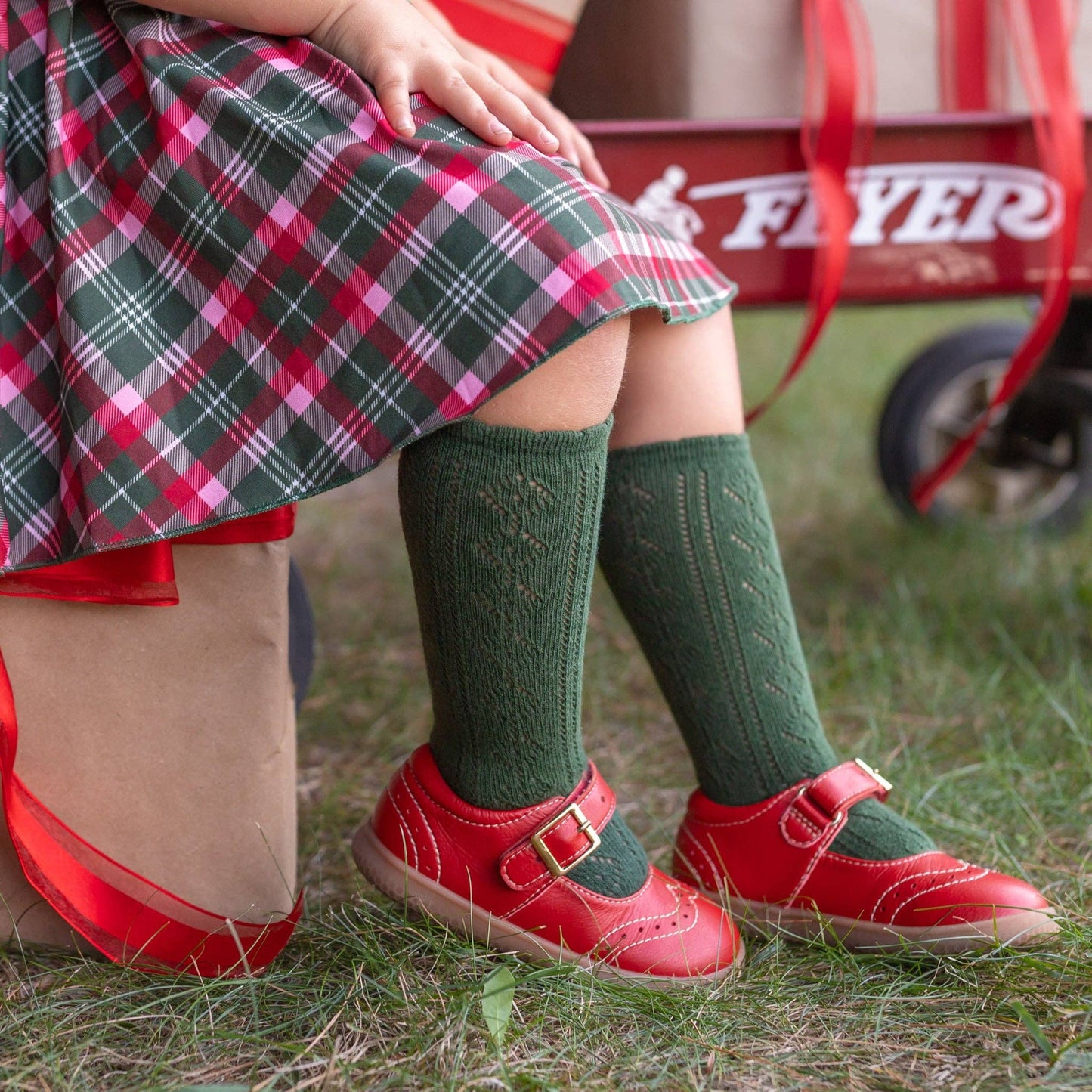 Child wearing green socks and red shoes sitting on grass next to a red wagon.