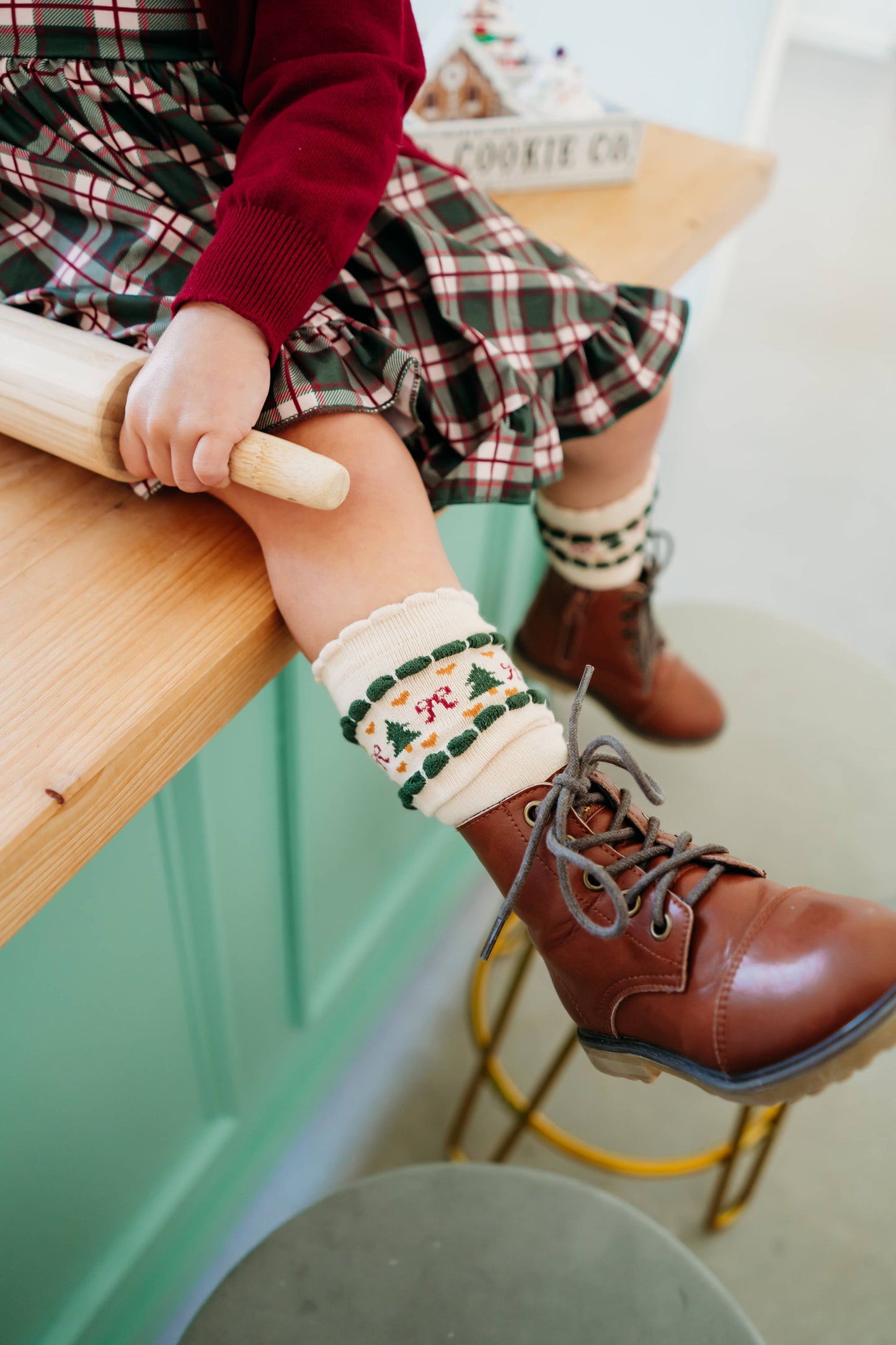 Person wearing a plaid skirt, red sweater, and brown boots with a Christmas-themed legwarmer in a kitchen.
