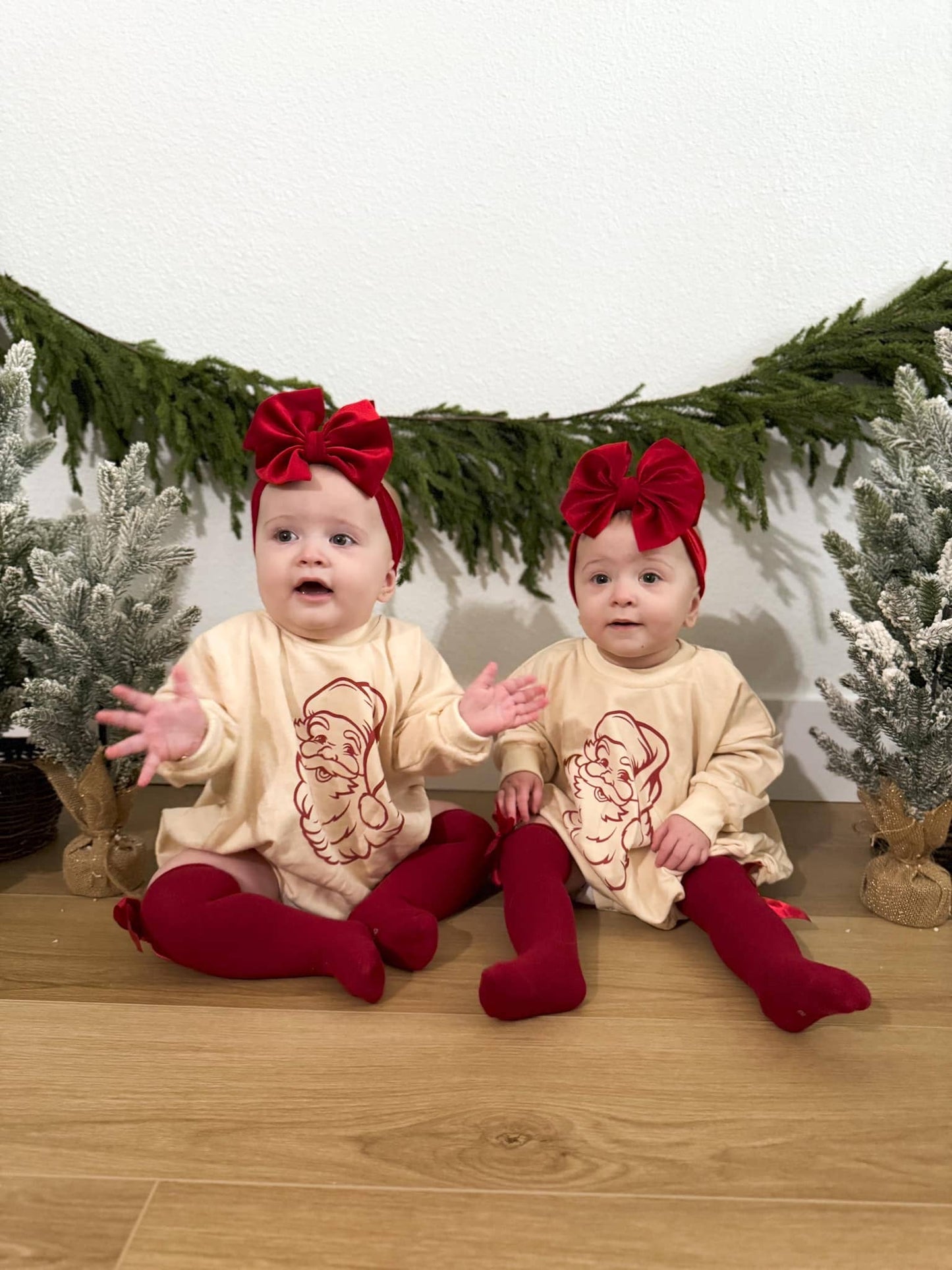 Two babies in matching outfits with red bows and stockings sitting on a wooden floor with Christmas decorations.