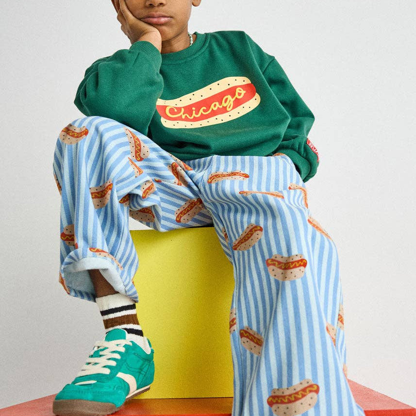 A child model wearing a green sweatshirt with a 'Chicago' graphic and light blue striped pants with a hot dog print, sitting on a yellow block atop a red platform against a light gray background.
