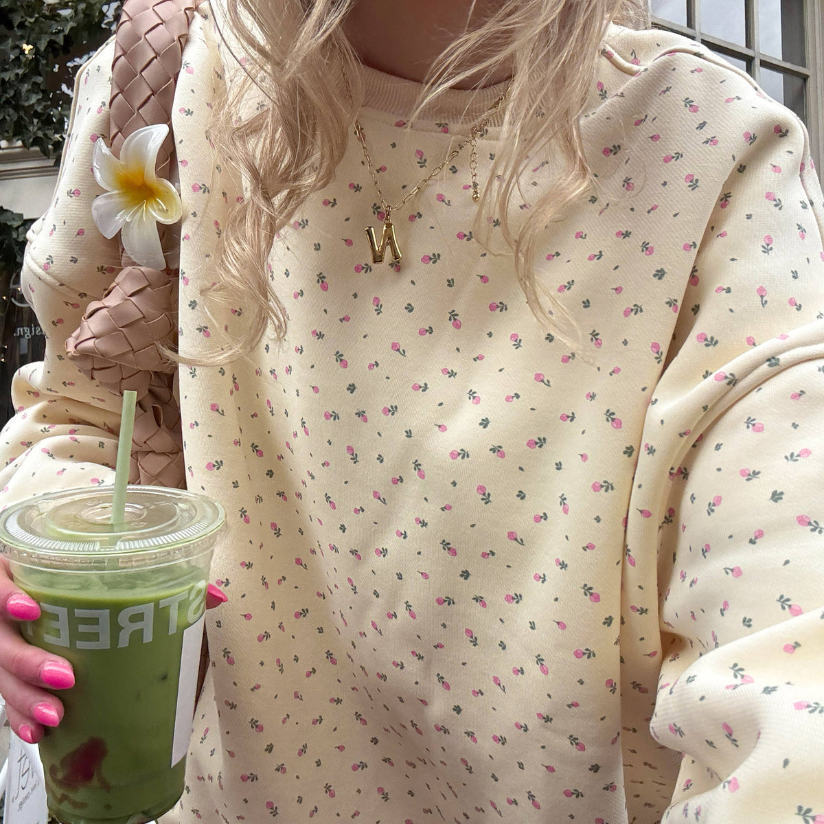 Person holding a green drink with a straw, wearing a light-colored floral-patterned hoodie.