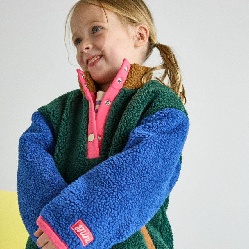 A young girl wearing a fleece pullover jacket with a green body, blue sleeves, and pink zipper trim, standing against a light gray wall.