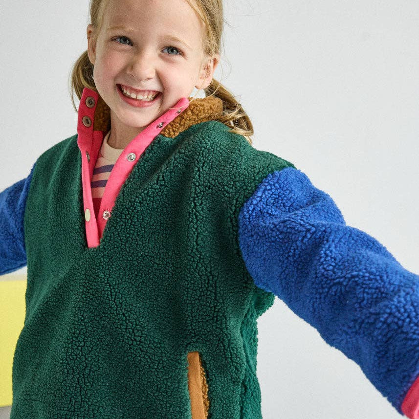 A young girl smiling and wearing a colorful fleece vest with a green body, blue sleeves, and a pink collar, paired with pink and blue gingham pants.