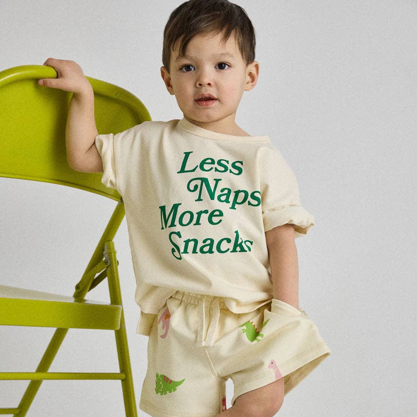 Child wearing a cream-colored outfit with green text and graphics, standing next to a green chair.