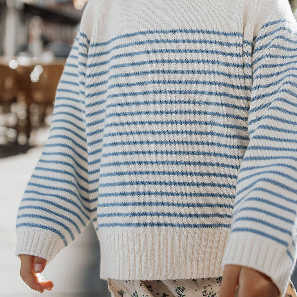 Child wearing a white sweater with blue stripes and floral pants outdoors.