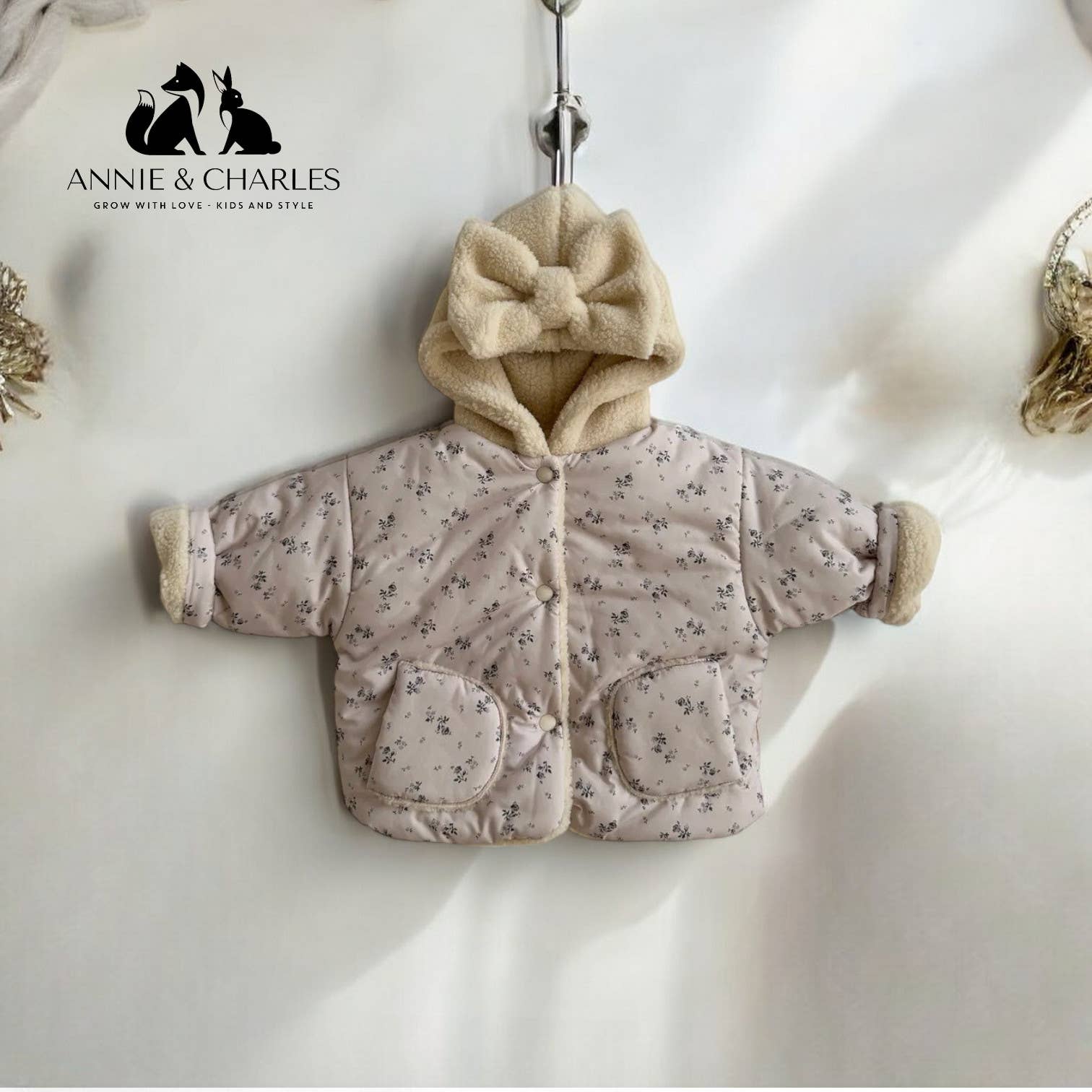 Baby jacket with floral pattern and bow on hood, hanging against a white background with 'Annie & Charles' branding.