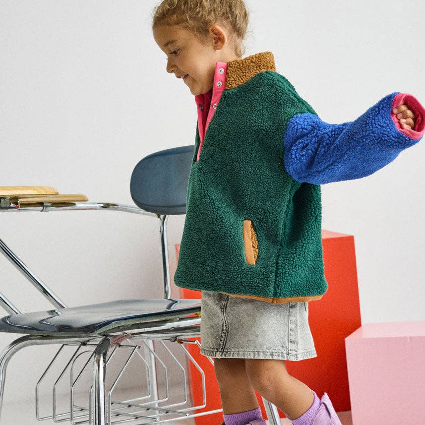 A child wearing a green sherpa fleece zip-up jacket with blue and pink sleeves, a denim skirt, and lavender suede boots, standing in a studio with chairs and colorful blocks.