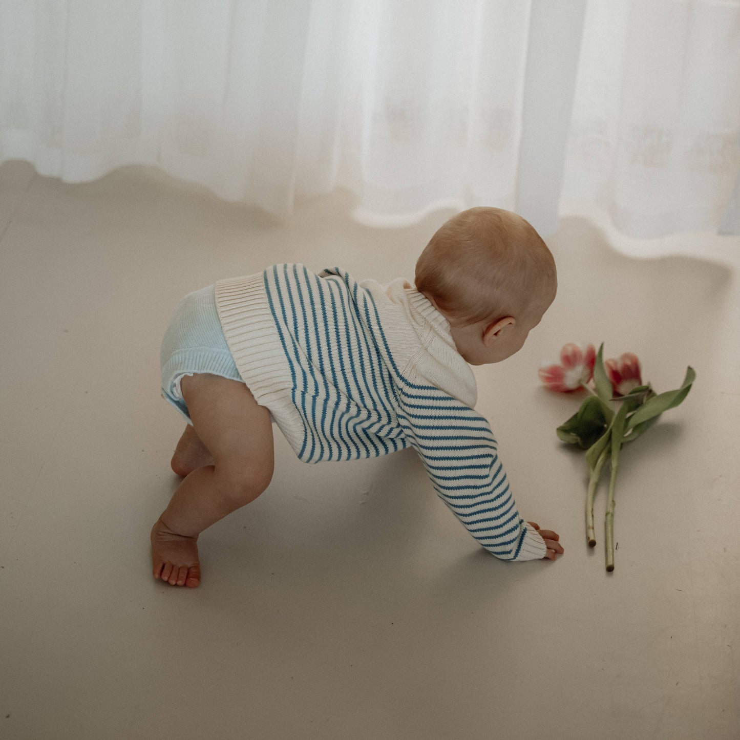Baby in a striped outfit crawling on a white surface with a flower nearby, white curtain in the background