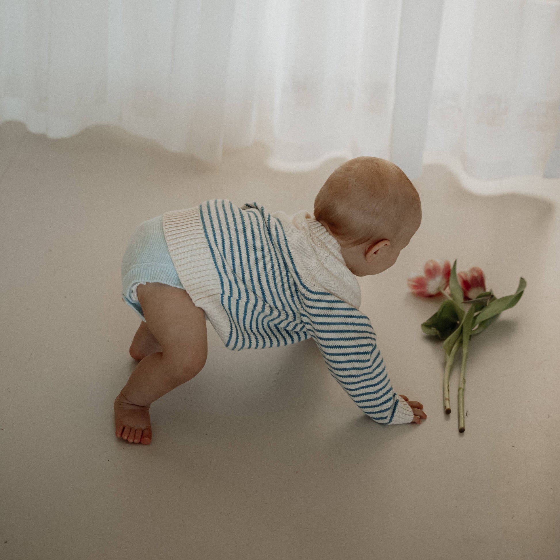 Baby in a striped outfit crawling on a white surface with a flower nearby, white curtain in the background