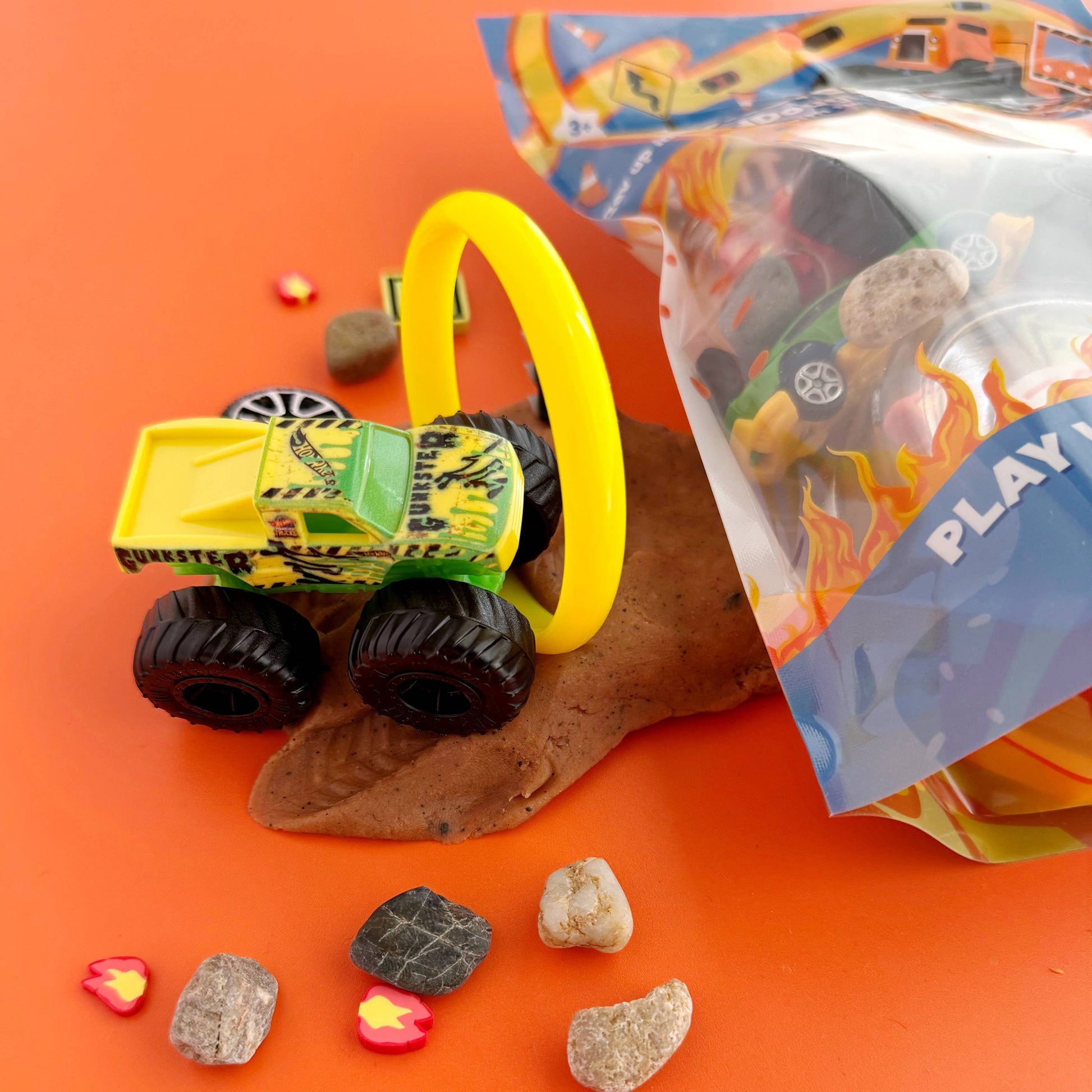Toy truck with a yellow handle on an orange surface, surrounded by small stones and a bag labeled 'Play'.