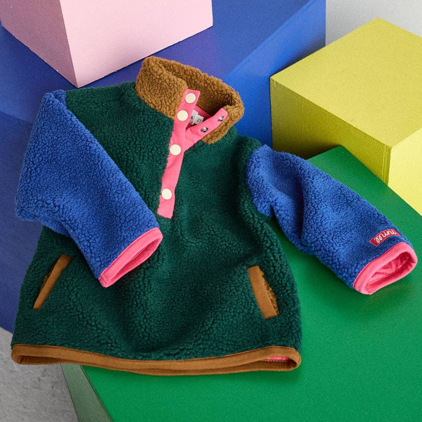 A children's fleece jacket laid flat on colorful geometric blocks. The jacket has a dark green body, bright blue sleeves, a brown collar, and a pink button placket with white buttons.