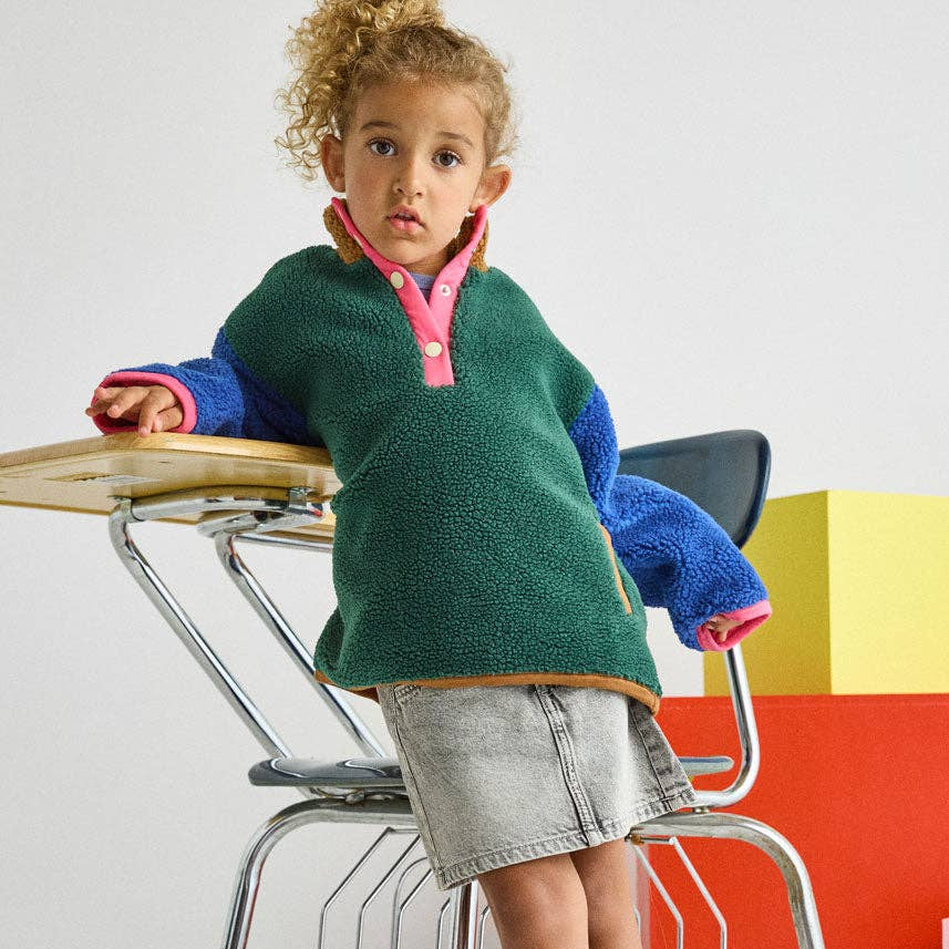 A young child wearing a green fleece pullover with blue and pink sleeves, a denim skirt, and purple boots, leaning against a school desk in a studio setting.