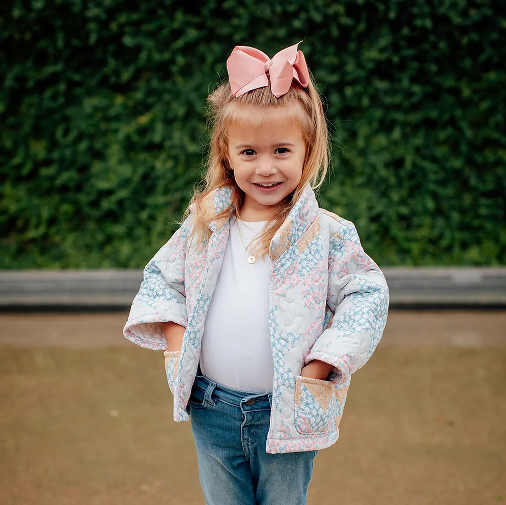 Young girl wearing a patterned jacket and pink bow, standing in front of green bushes.