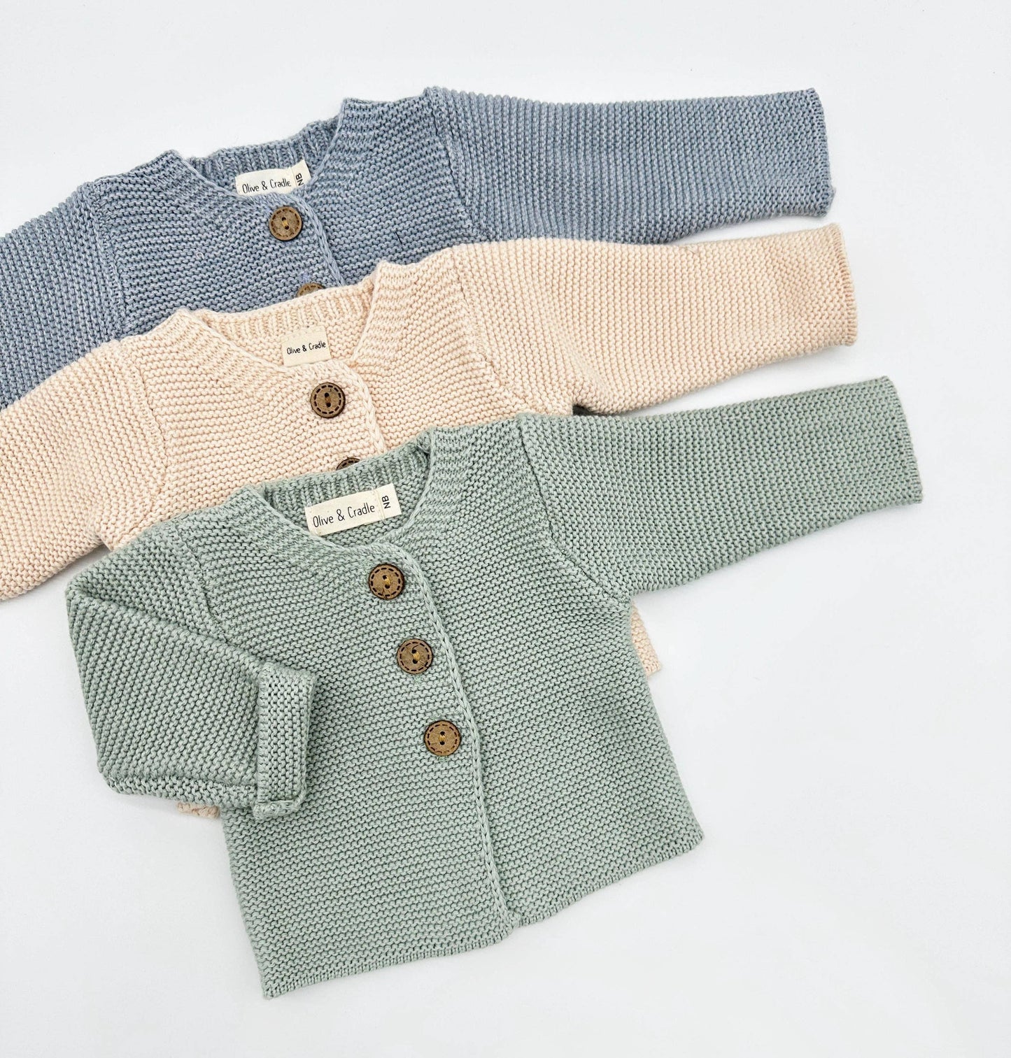 Three knitted baby cardigans in blue, beige, and green on a light gray background.
