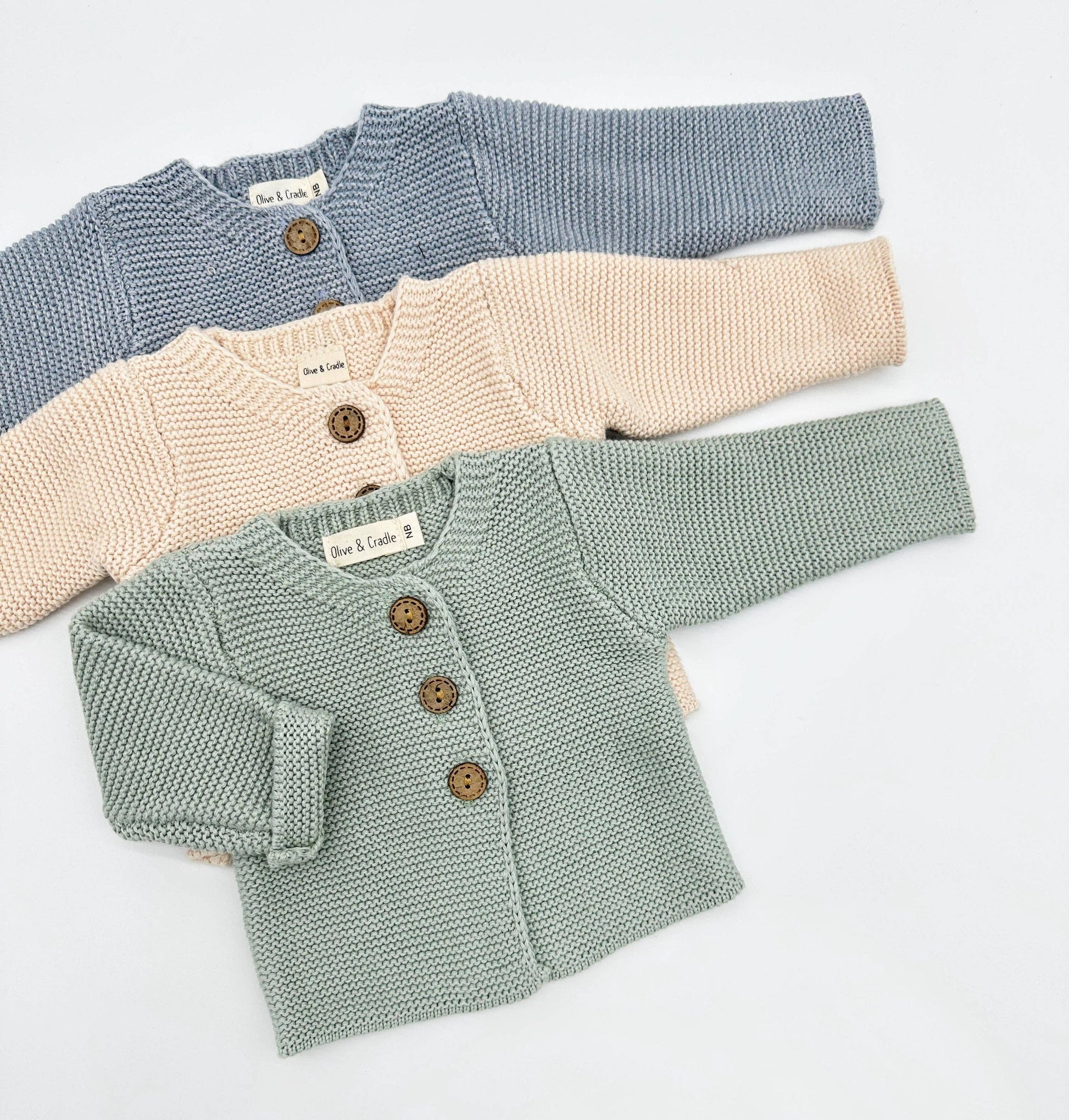 Three knitted baby cardigans in blue, beige, and green on a light gray background.