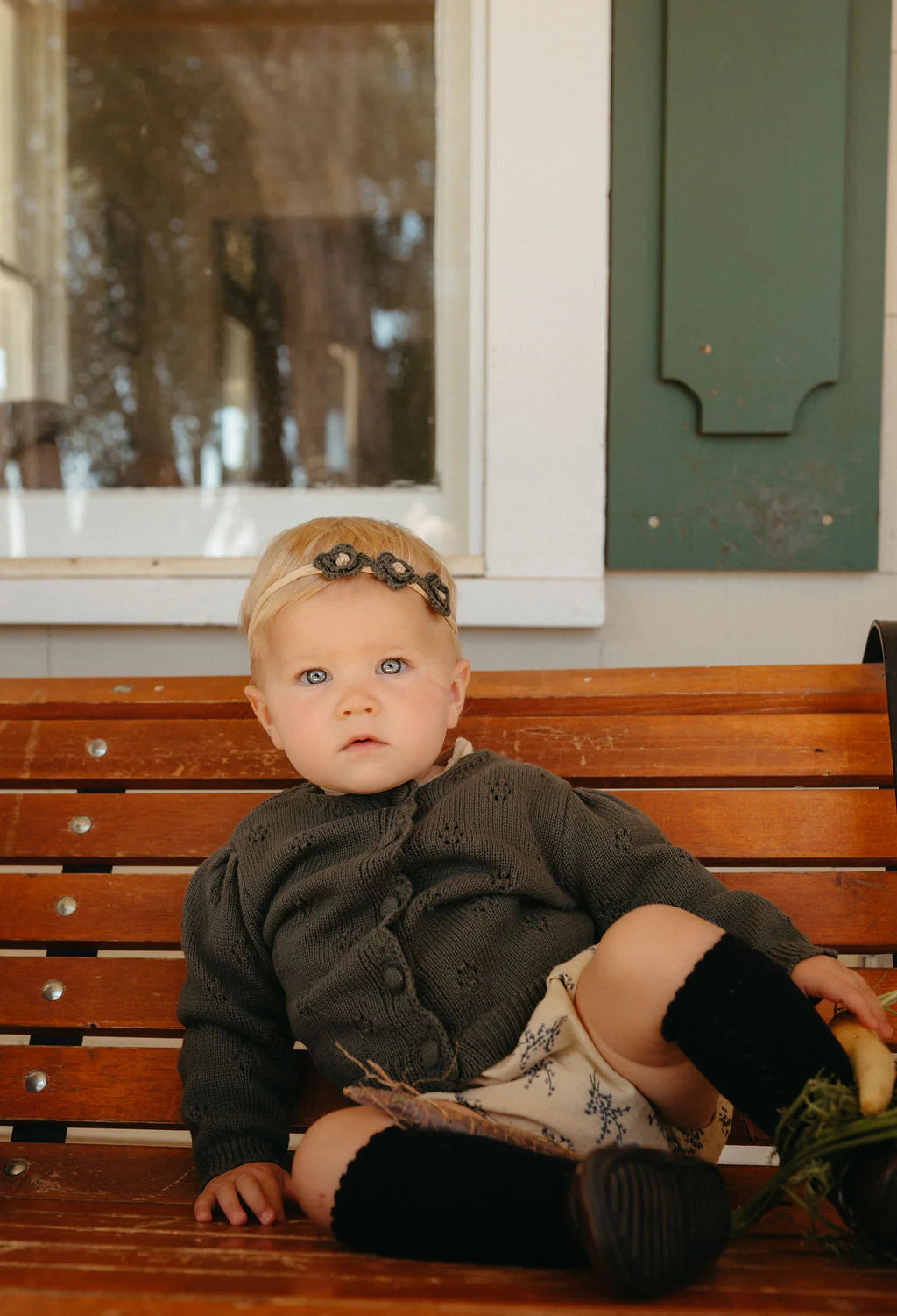 Child sitting on a wooden bench wearing a dark green cardigan and black leggings.