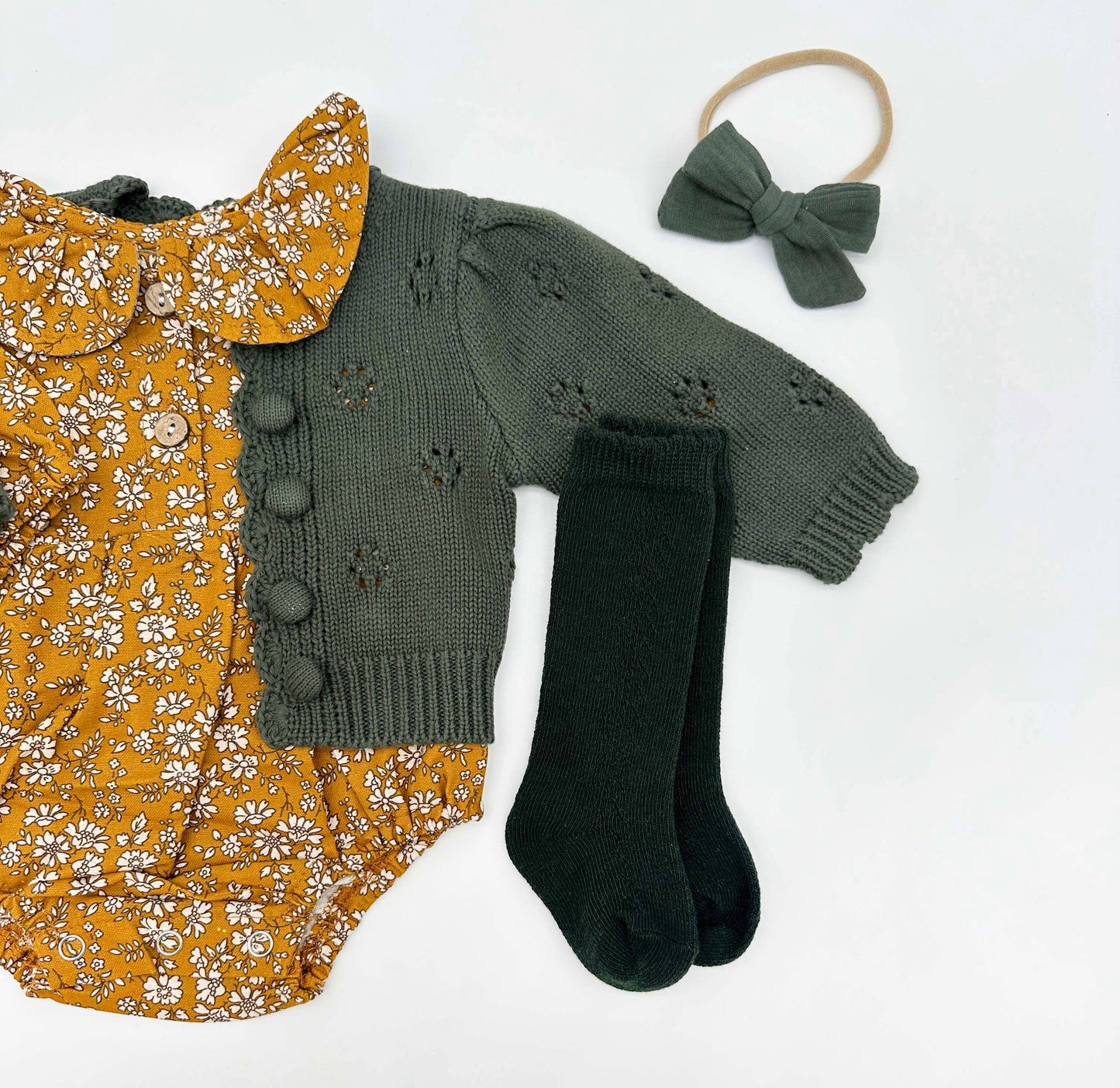 Baby outfit with yellow floral dress, green knitted cardigan, black socks, and headband on a white background