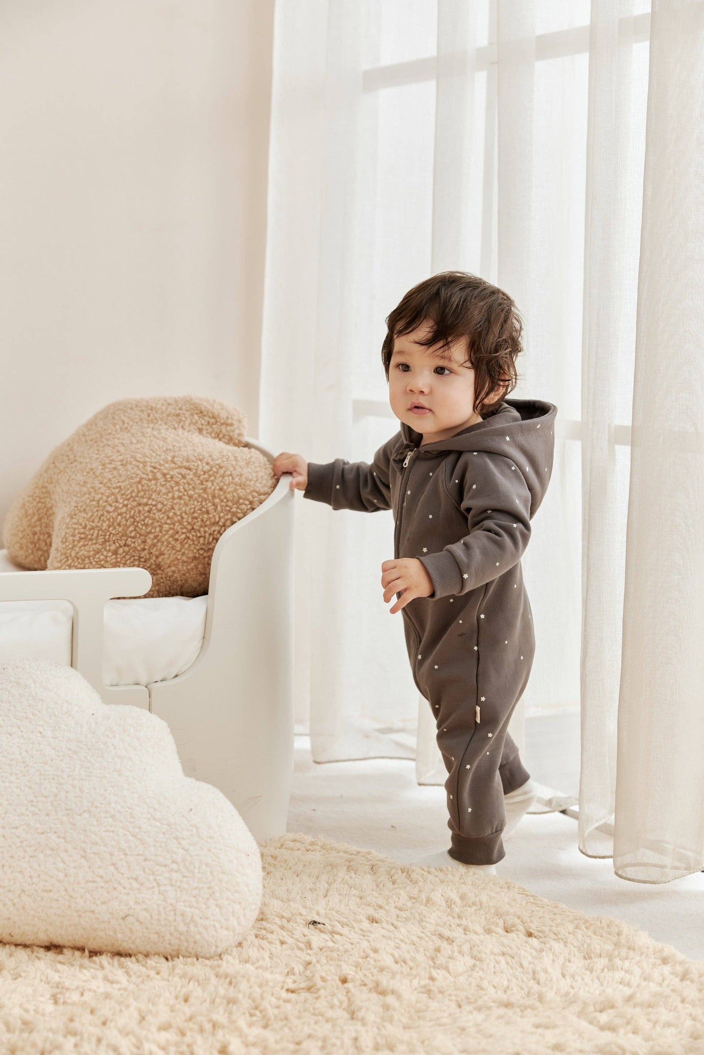 Child in a gray onesie standing in a bright room with white furniture and beige rug.