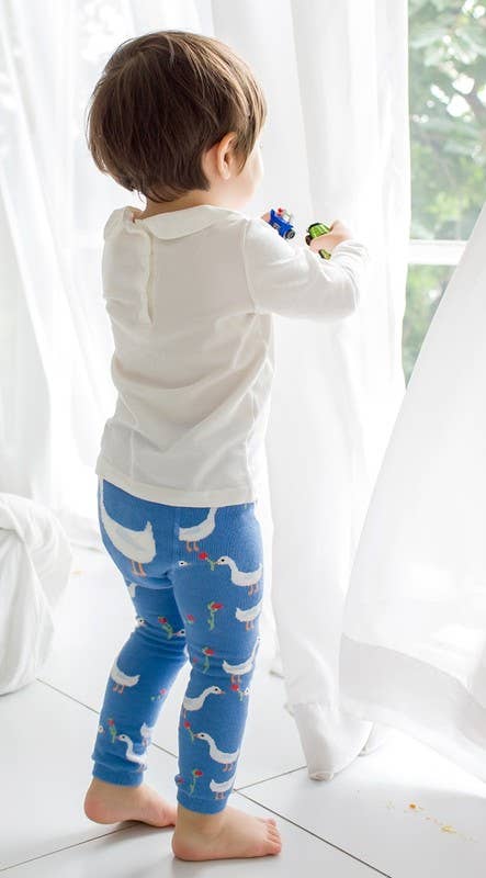 Child wearing a white long-sleeve shirt and blue leggings with bird pattern in a bright room.