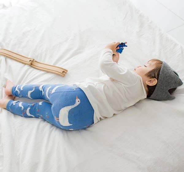 Child wearing blue leggings with bird pattern and gray hat lying on a white bed.