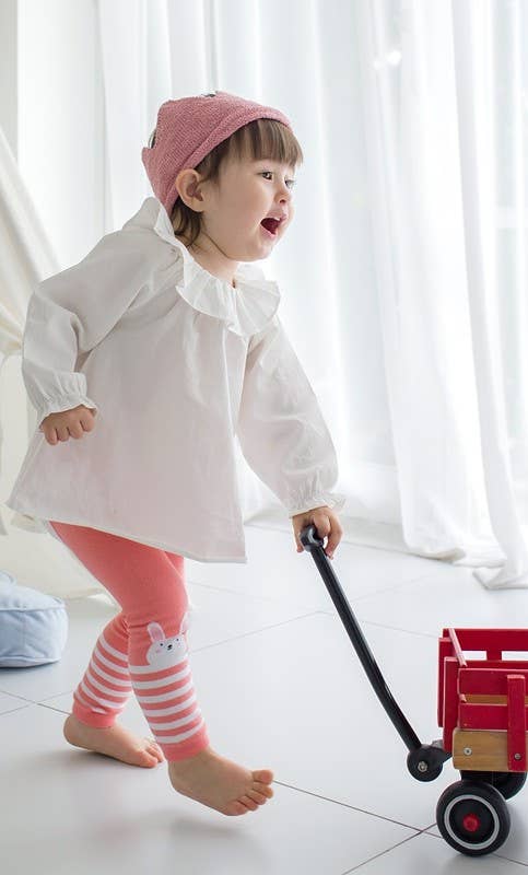 Child in a white coat and pink pants with a red toy wagon on a light background