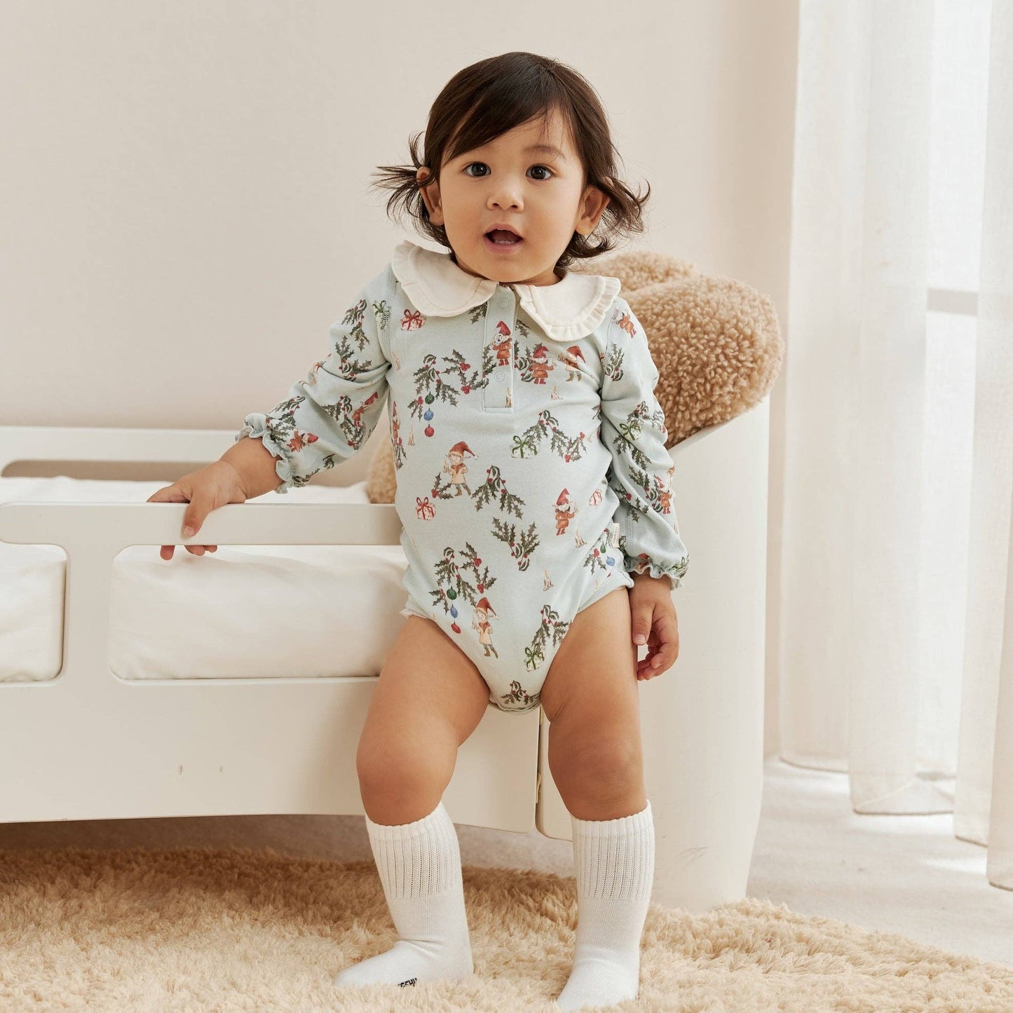 Child wearing a floral onesie standing in a room with a white couch and light-colored walls.