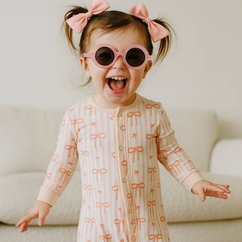 Child wearing a onesie with bow pattern and sunglasses, standing in a room.