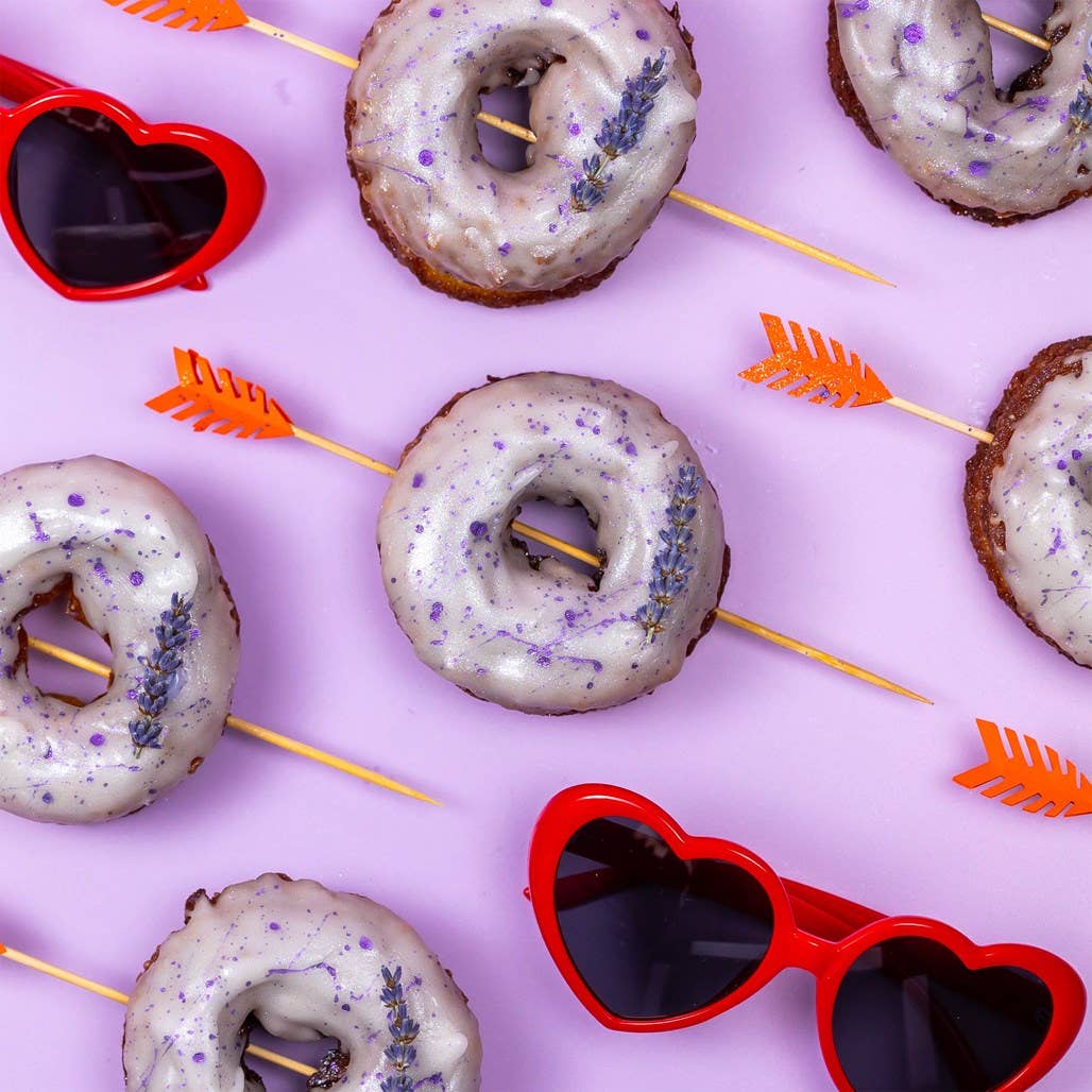 Donuts with white glaze and purple sprinkles on toothpicks with red heart sunglasses and orange arrows on a pink background.
