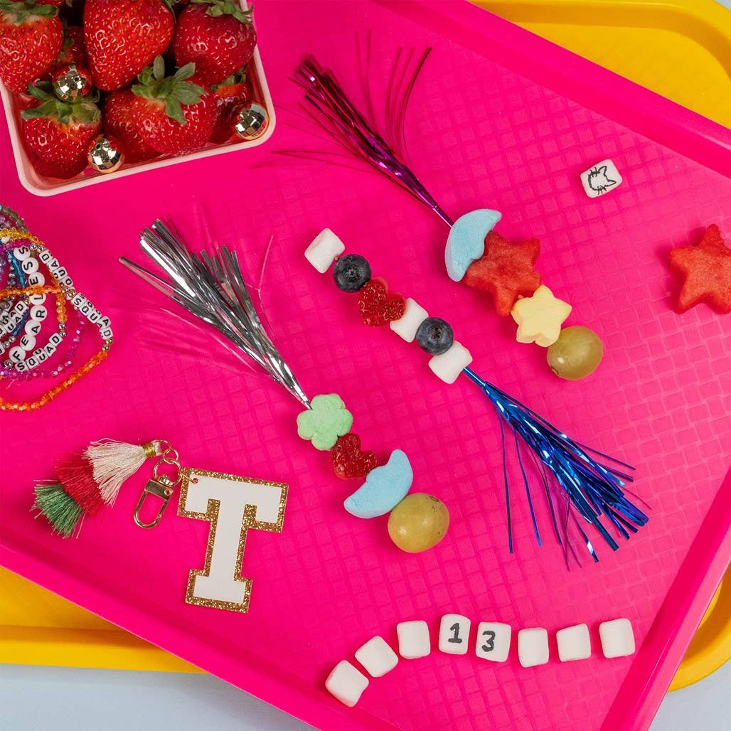 Colorful tray with decorative items including a letter 'T', tassels, and small fruits on a pink background.