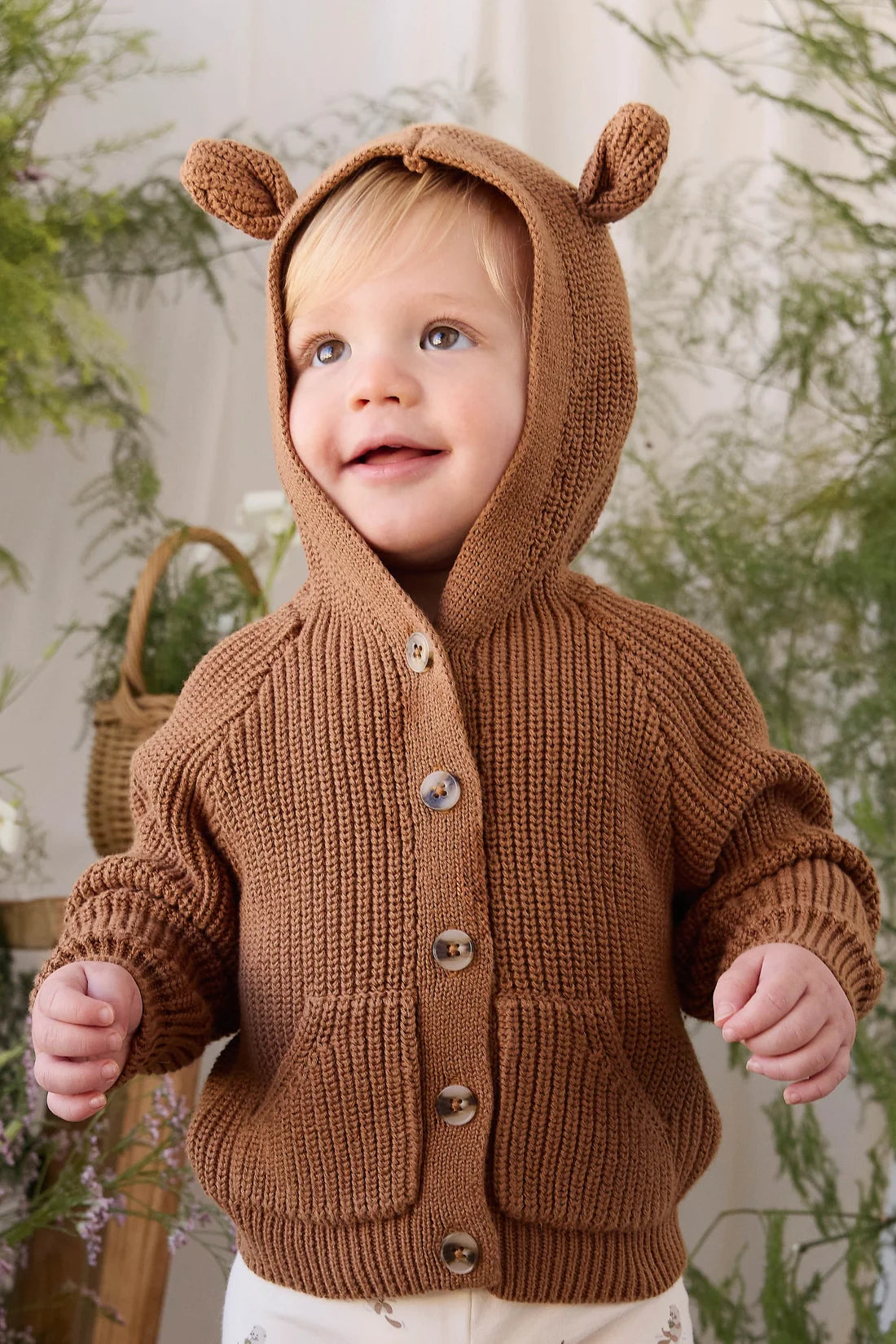 A toddler wearing a brown knit hooded cardigan with bear ears on the hood and front buttons, standing in a softly decorated indoor setting with plants and a basket.