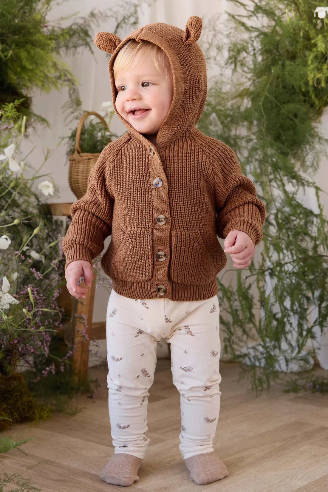 A smiling toddler standing indoors wearing a brown ribbed knit hooded cardigan with bear ears and cream pants with a small animal print.