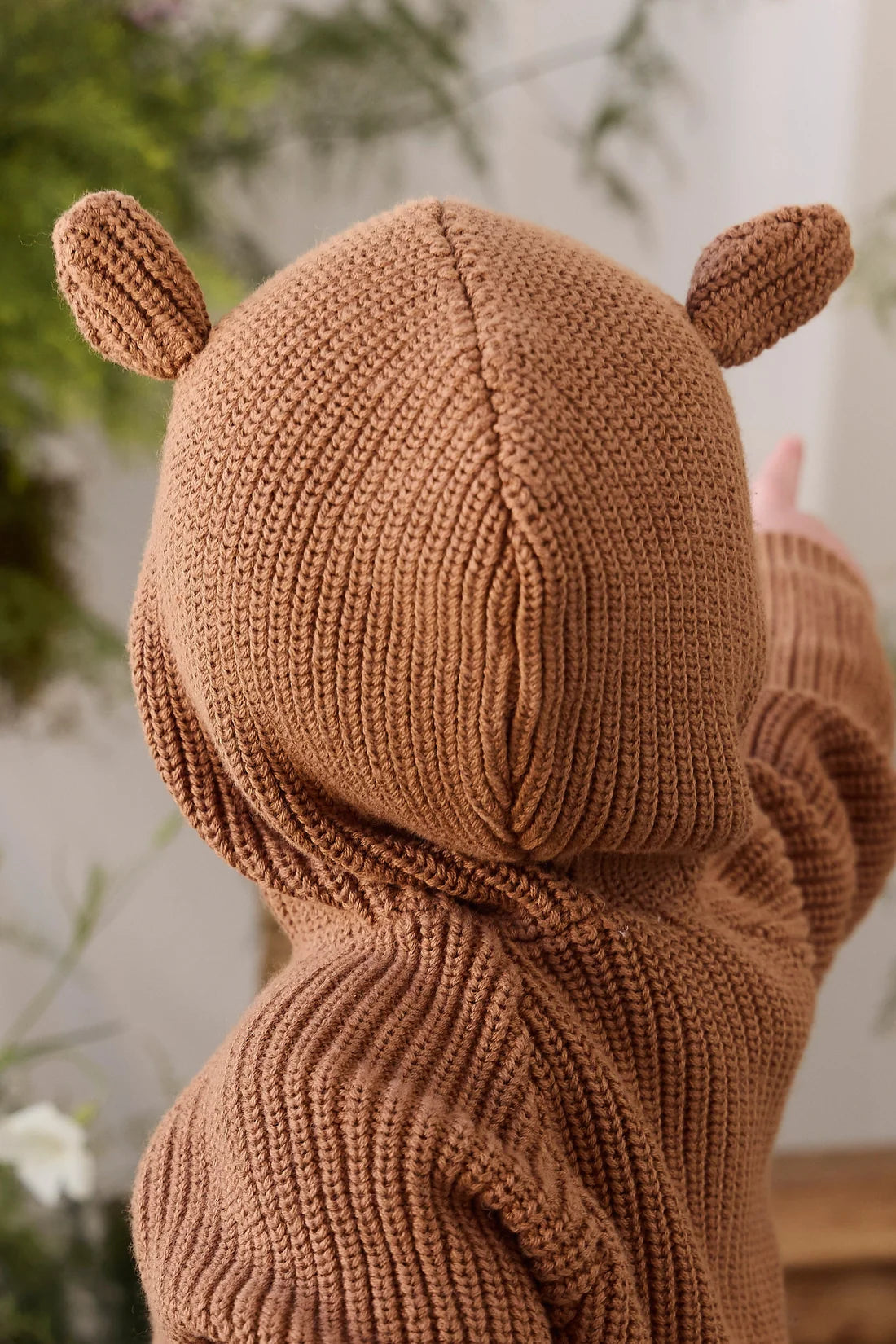 A close-up of a child wearing a cozy, ribbed knit hooded sweater in a warm brown color, featuring small ear details on the hood. The photo is taken from behind, showing the texture of the knit fabric against a softly blurred indoor background with green plants.