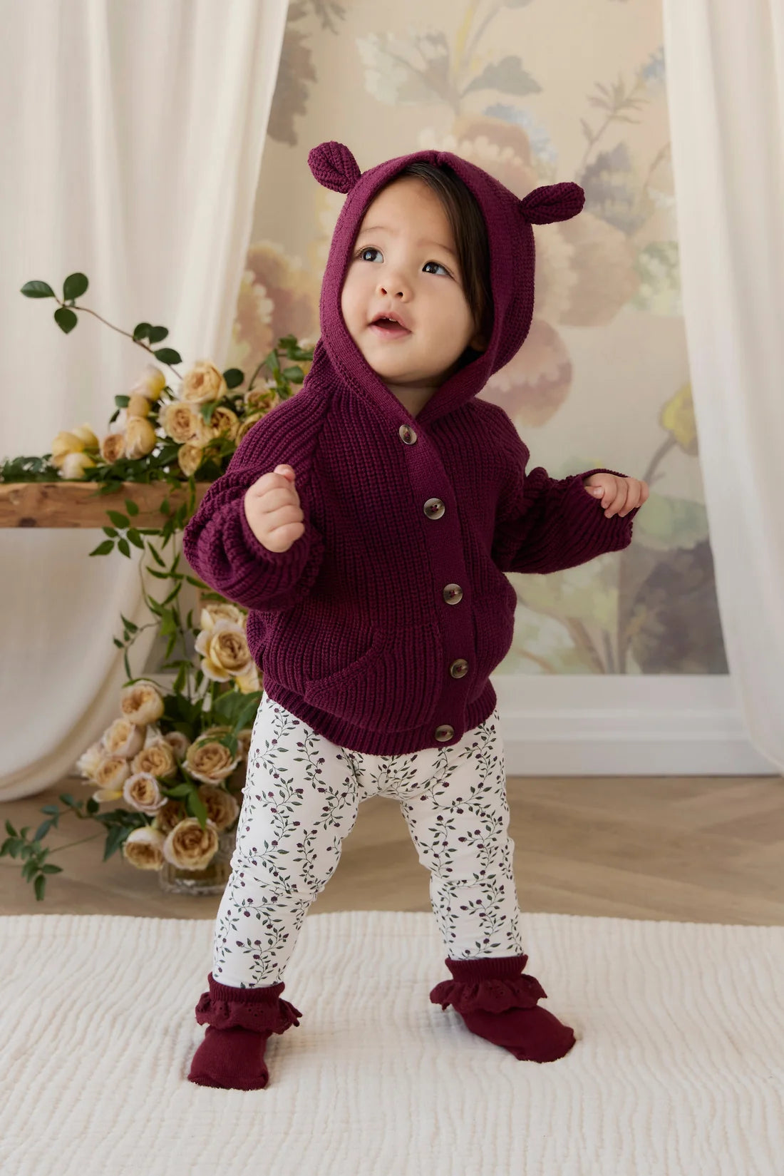 A toddler standing indoors wearing a chunky knit burgundy hooded cardigan with bear ears, white patterned leggings, and matching burgundy booties.