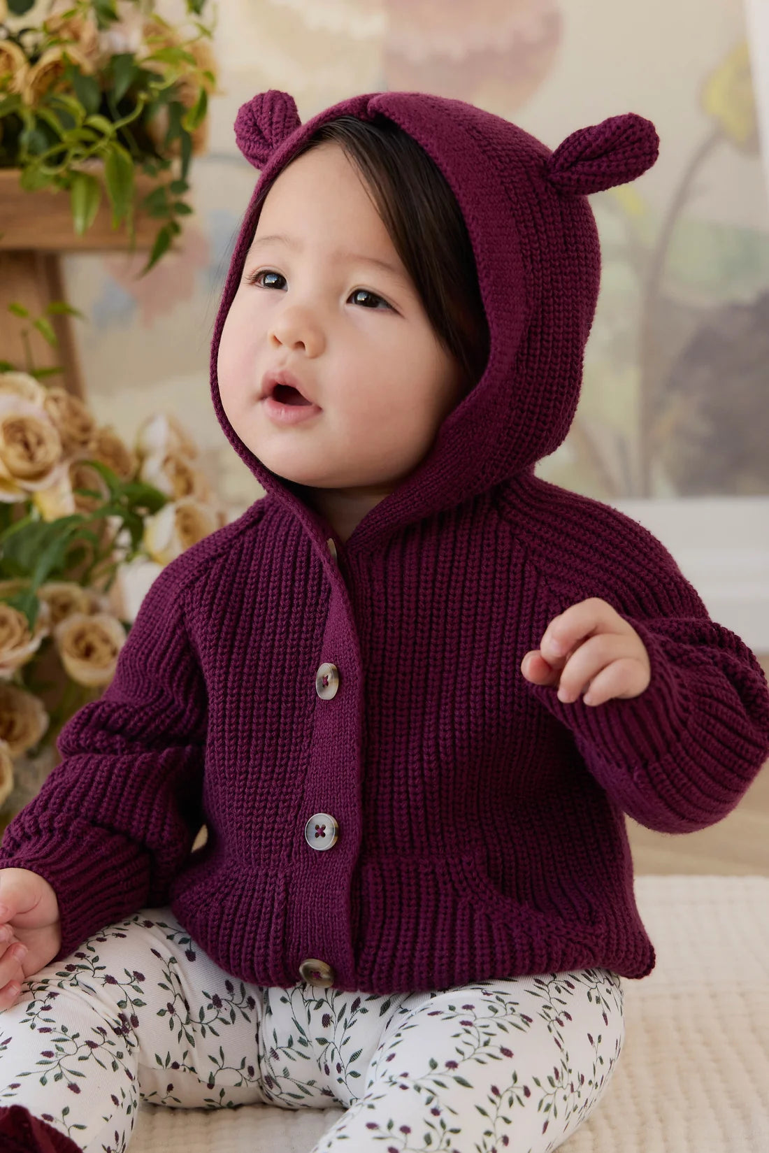 A baby wearing a knitted burgundy hooded cardigan with bear ears and white patterned pants, sitting in a cozy indoor setting with flowers in the background.