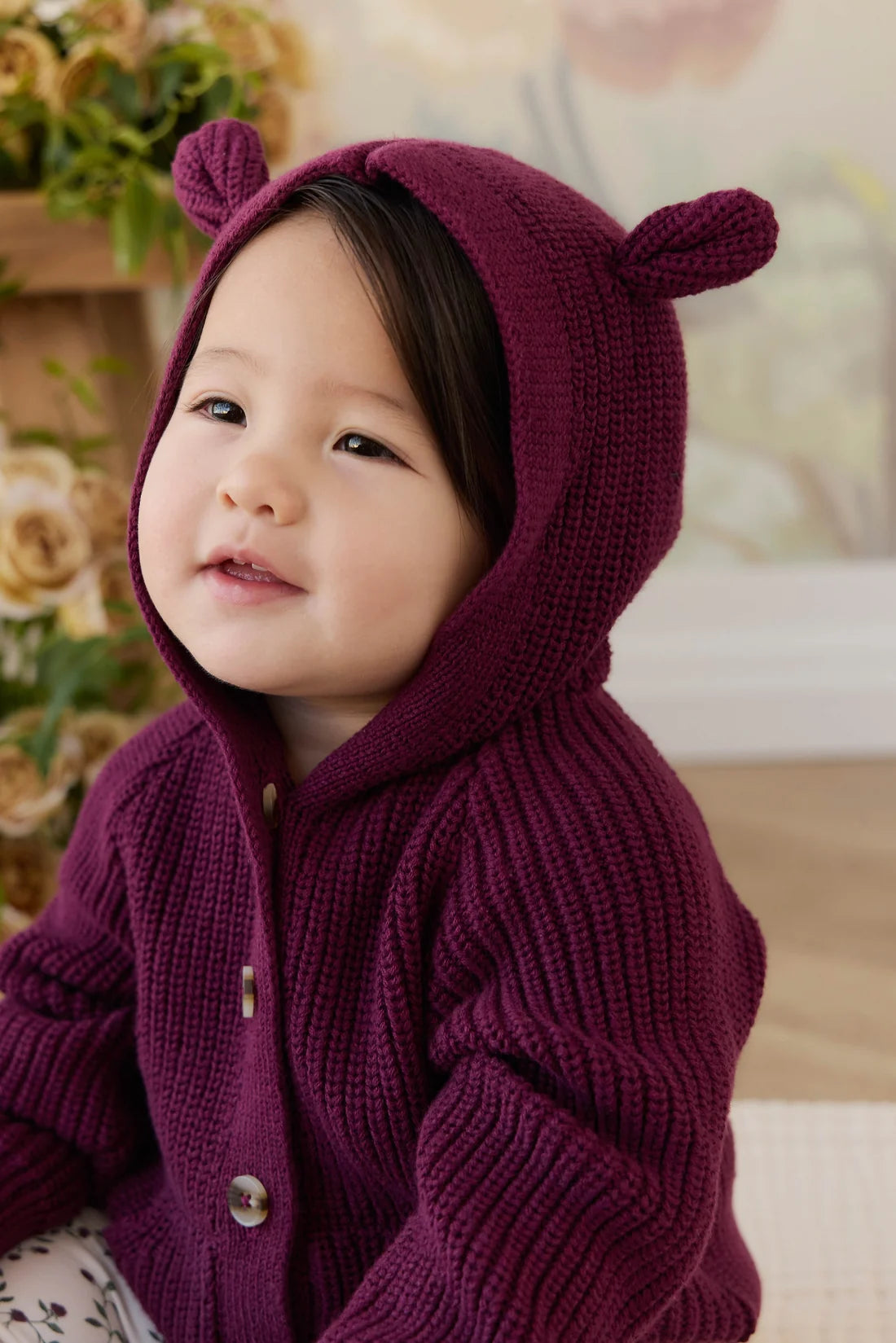 A baby wearing a chunky knit burgundy cardigan with a hood featuring bear ears, photographed in a soft indoor setting.