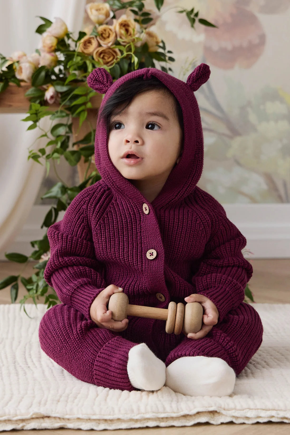 A baby wearing a cozy, chunky-knit burgundy hooded jumpsuit with bear ears sits on a cream textured rug, holding a wooden toy.