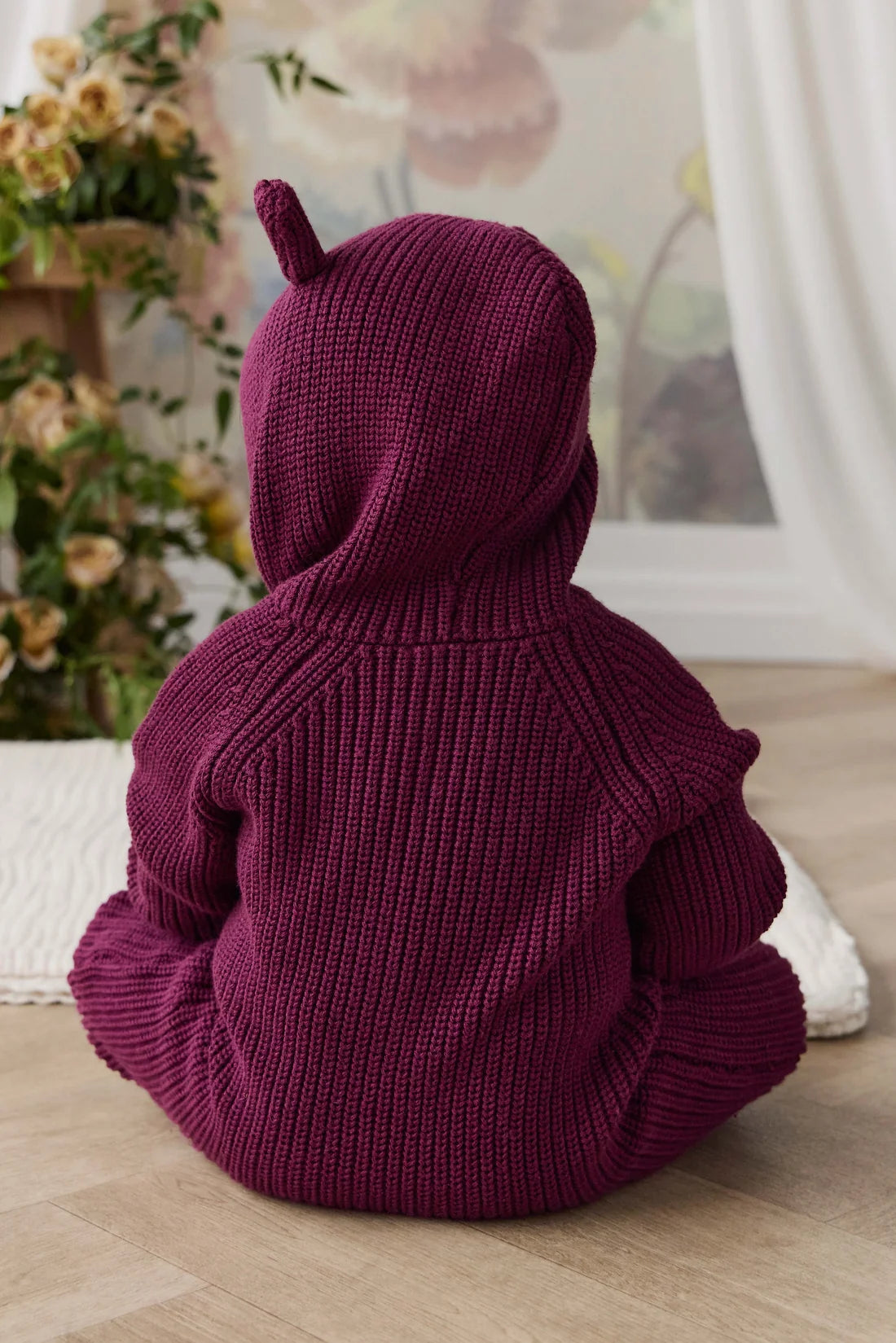 A baby wearing a deep burgundy ribbed knit hooded sweater, seen from behind, sitting on a light wood floor in a softly decorated room.