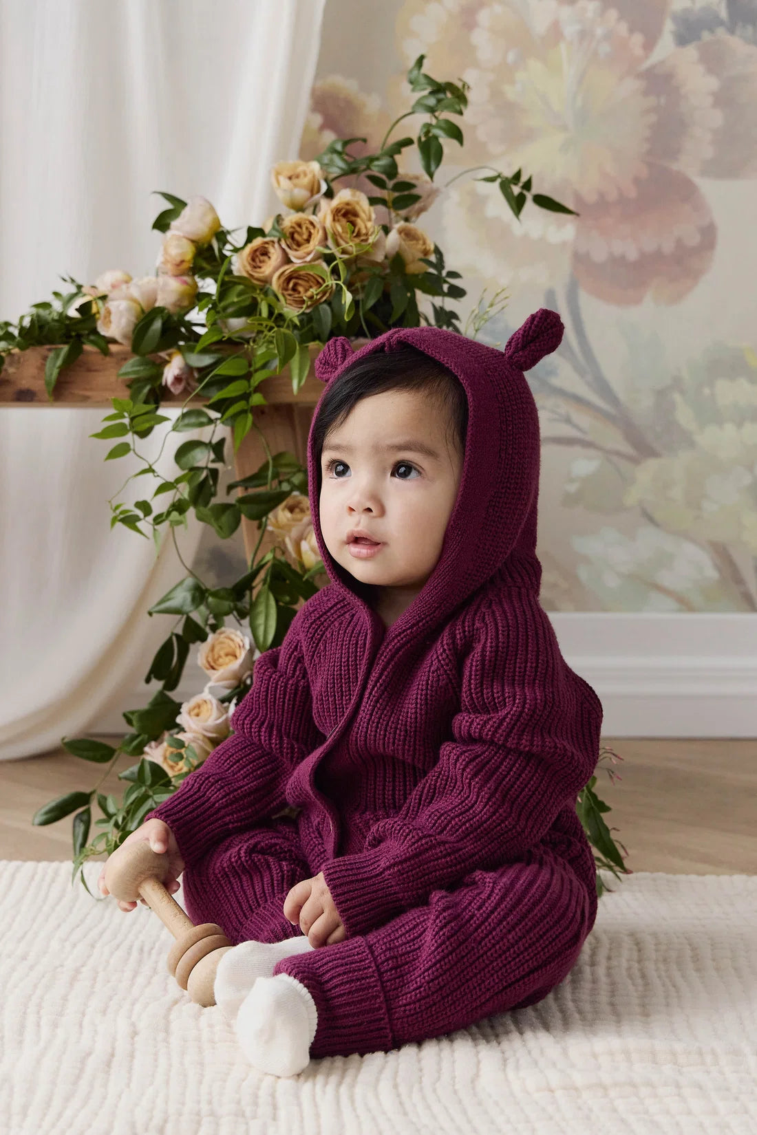 A baby wearing a cozy, deep purple ribbed knit hooded romper with bear ears sits on a cream textured rug. The background features a soft floral wall mural, draped white curtains, and a decorative arrangement of green foliage and cream roses.