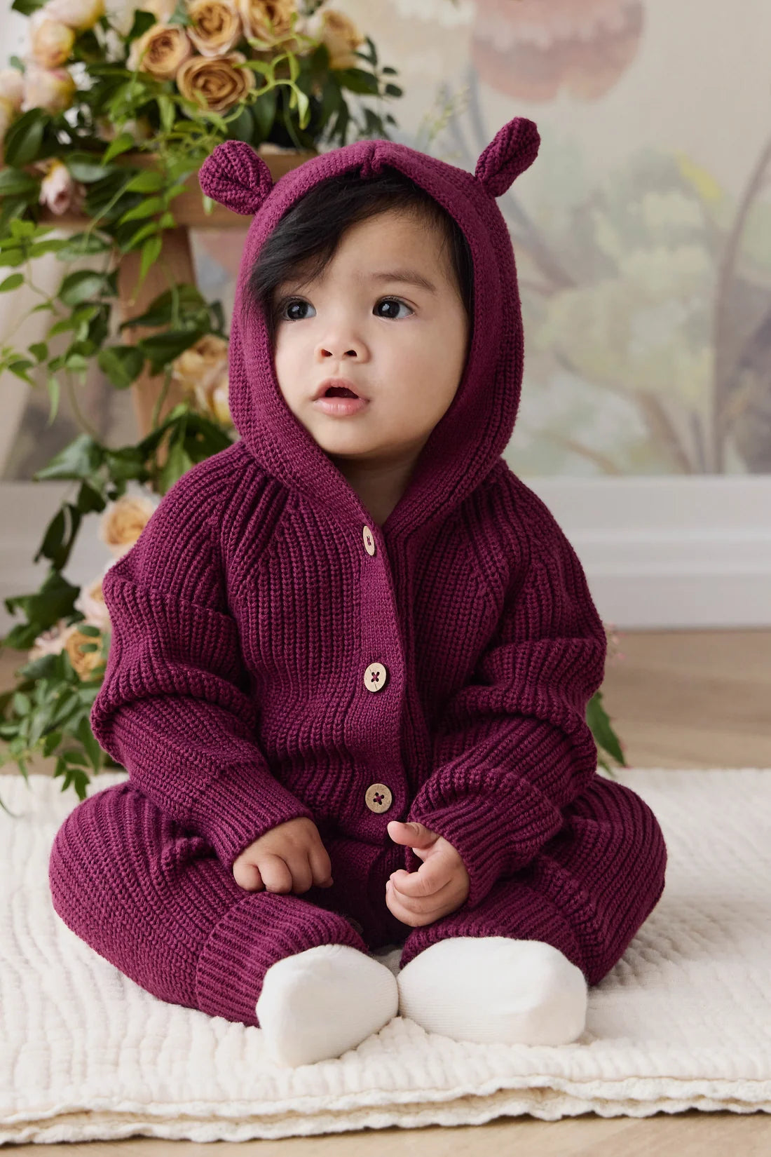 A baby wearing a chunky knit burgundy hooded jumpsuit with bear ears on the hood and white socks, sitting on a white textured blanket.