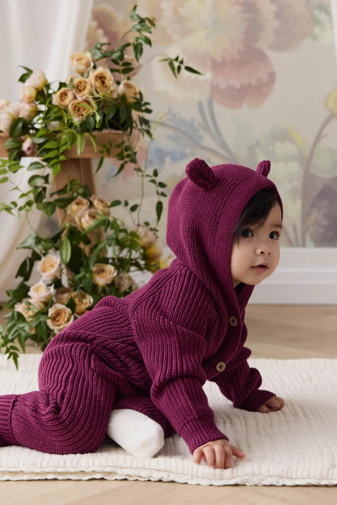A baby crawling on a cream textured rug wearing a deep purple ribbed knit hooded jumpsuit with bear ears on the hood, set in a warm room with floral wallpaper and a flower arrangement.